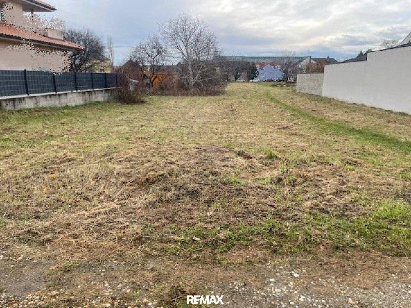 Weitläufiges, unbebautes Grundstück mit Grasfläche, begrenzt durch einen Zaun und eine Mauer.