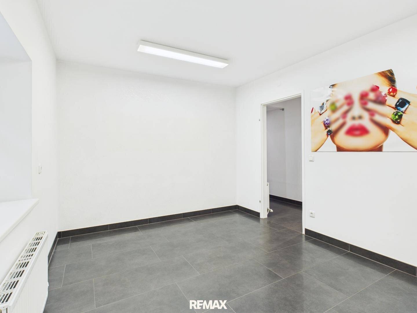Leerer, heller Raum mit weißen Wänden, grauem Fliesenboden und einem dekorativen Wandbild, ideal als Büro.