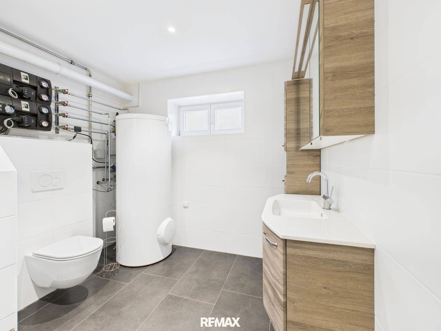 Badezimmer mit Toilette, Waschbecken, Warmwasserspeicher und sichtbaren Rohrleitungen, ausgestattet mit Fenster.