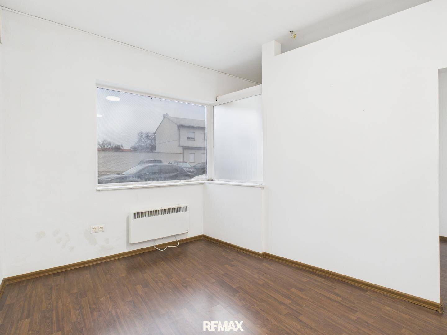 Heller Büroraum mit Laminatboden und einem großen Fenster, das viel Tageslicht hereinlässt.