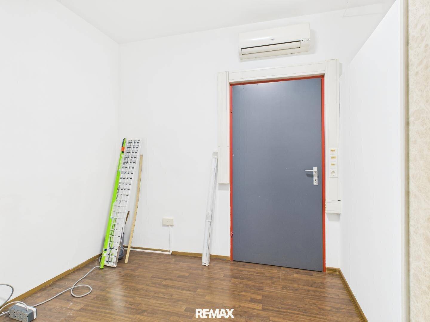 Kleiner Büroraum mit Laminatboden, weißen Wänden und einer grauen Tür, bereit zur individuellen Gestaltung.