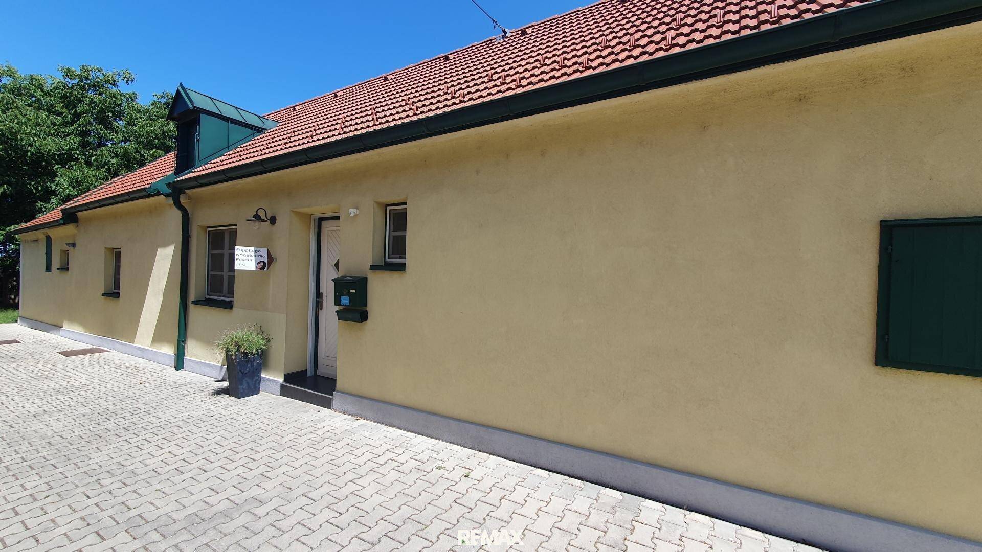 Seitenansicht des Gebäudes mit gelber Fassade, grünen Fensterläden und gepflastertem Zugangsbereich unter blauem Himmel.