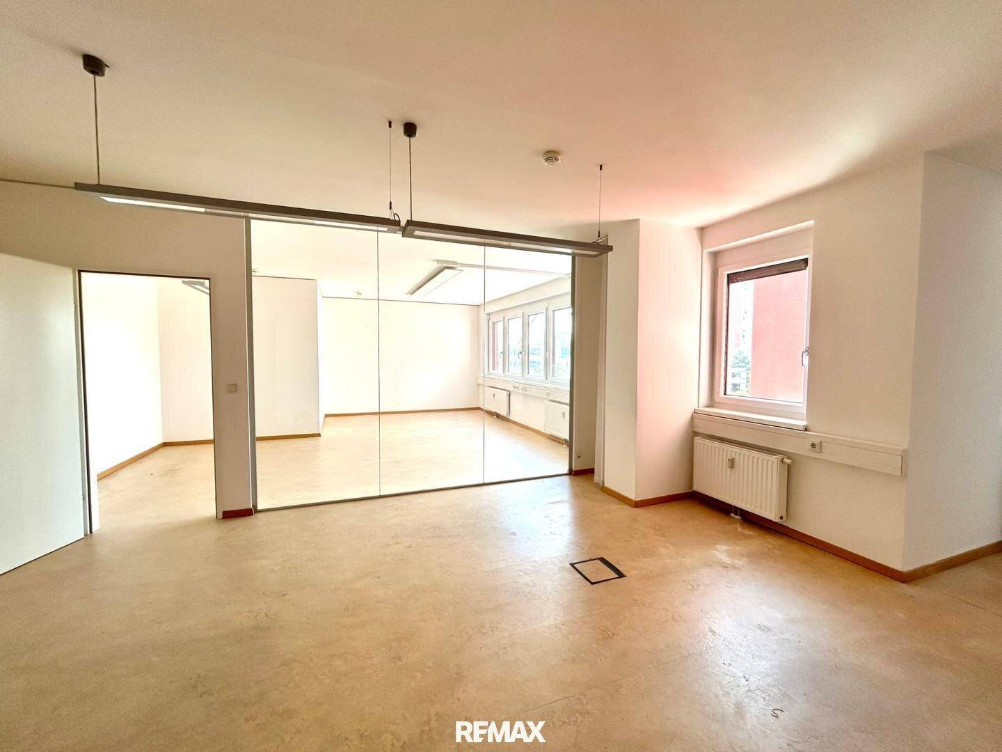 Helles Büro mit großen Fenstern, Vinylboden und einer Glaswand, die einen weiteren Raum abtrennt.