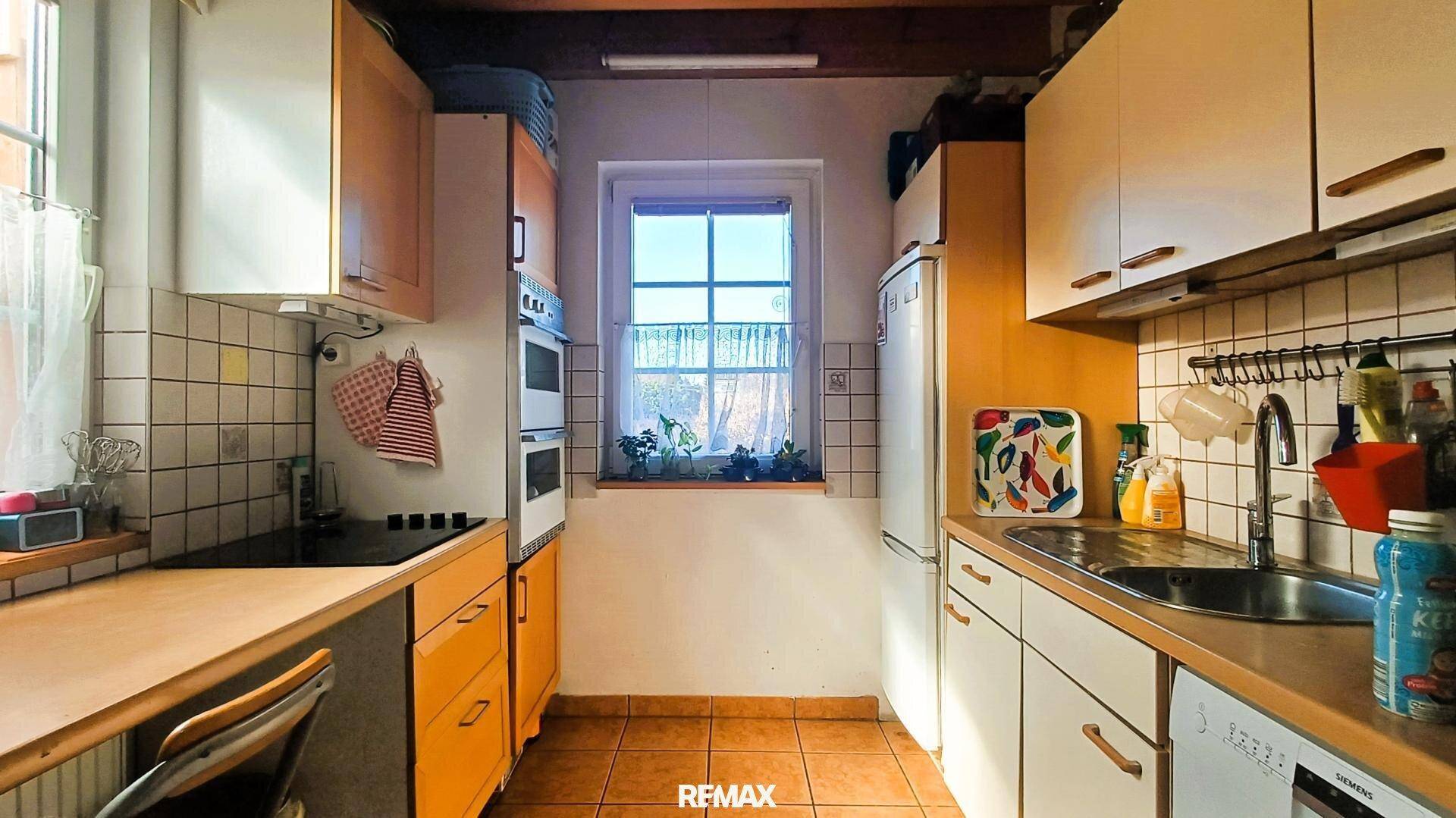 Funktionale Küche mit Holzschränken, Fliesenboden und einem Fenster, das viel Tageslicht hereinlässt.