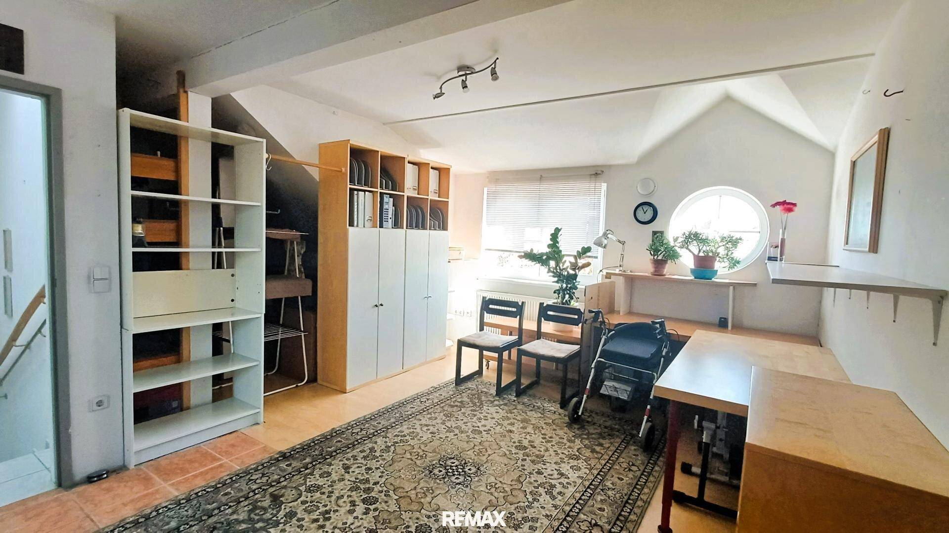 Dachgeschosszimmer mit schrägen Wänden, Holzboden und einem runden Fenster, als Arbeitsbereich genutzt.