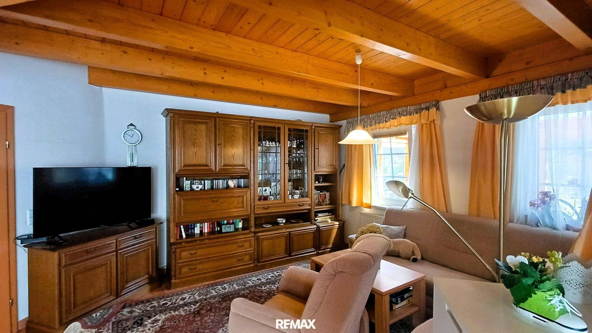 Wohnzimmer mit sichtbaren Holzbalken an der Decke, Teppichboden und traditionellen Holzmöbeln.