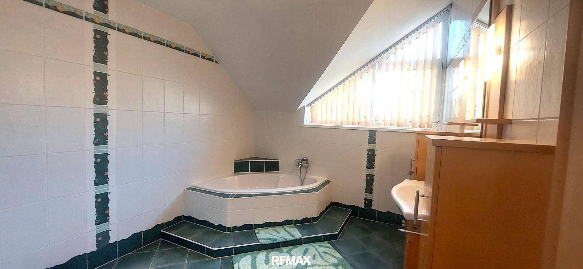 Badezimmer mit Eckbadewanne, grünen und weißen Fliesen sowie einem Fenster unter der Dachschräge.