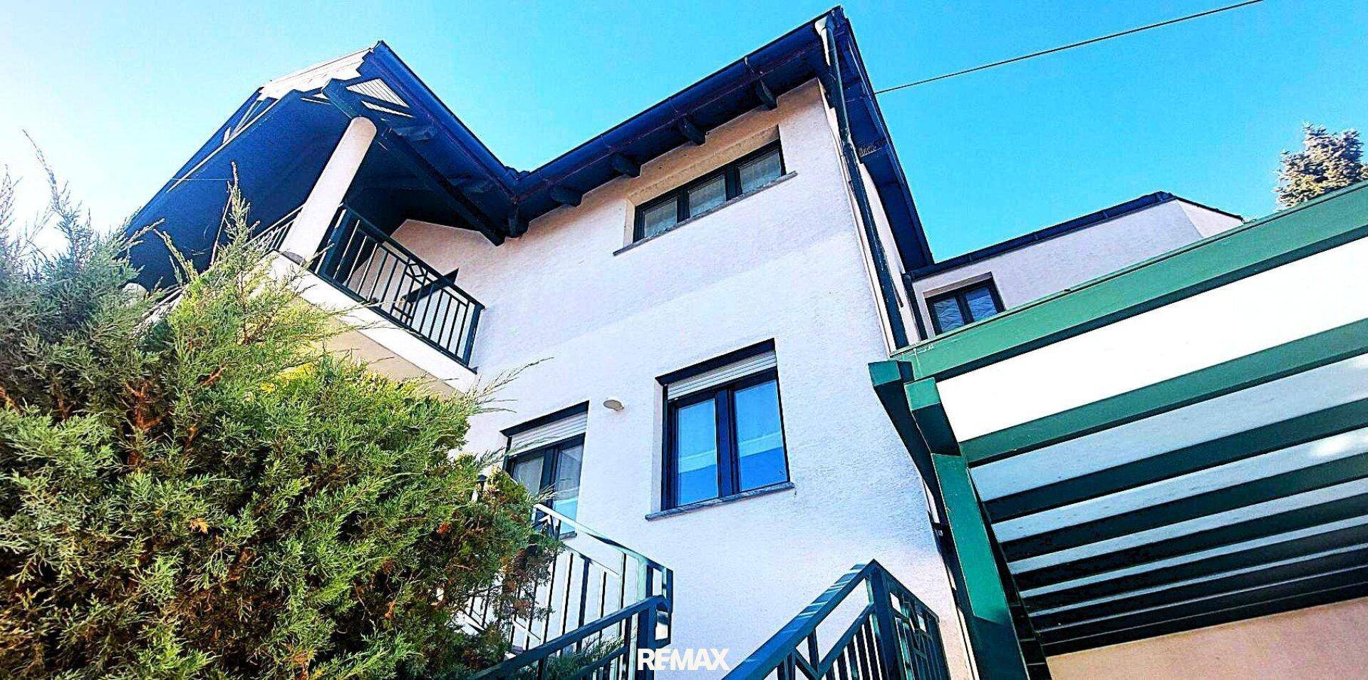 Ansicht des Hauses mit Balkon, dunklen Fensterrahmen und einem grünen Carport unter strahlend blauem Himmel.