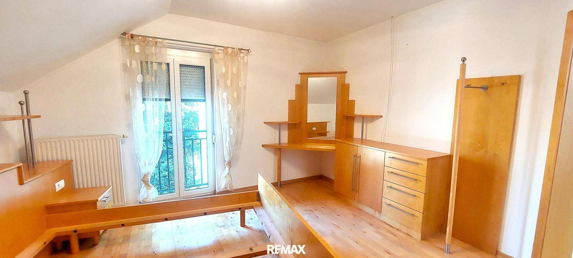Schlafzimmer mit Holzbettrahmen, Schminktisch und Fenster mit Vorhängen unter der Dachschräge.