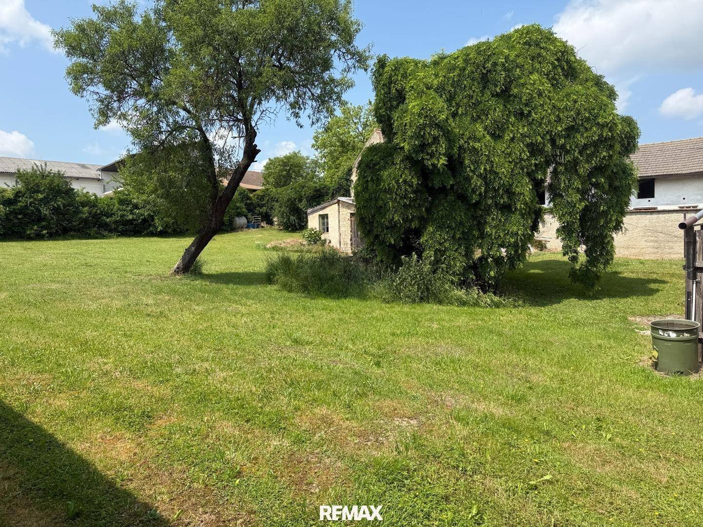 Eine weite Rasenfläche mit einigen Bäumen und einem kleinen Gebäude im Hintergrund unter blauem Himmel.