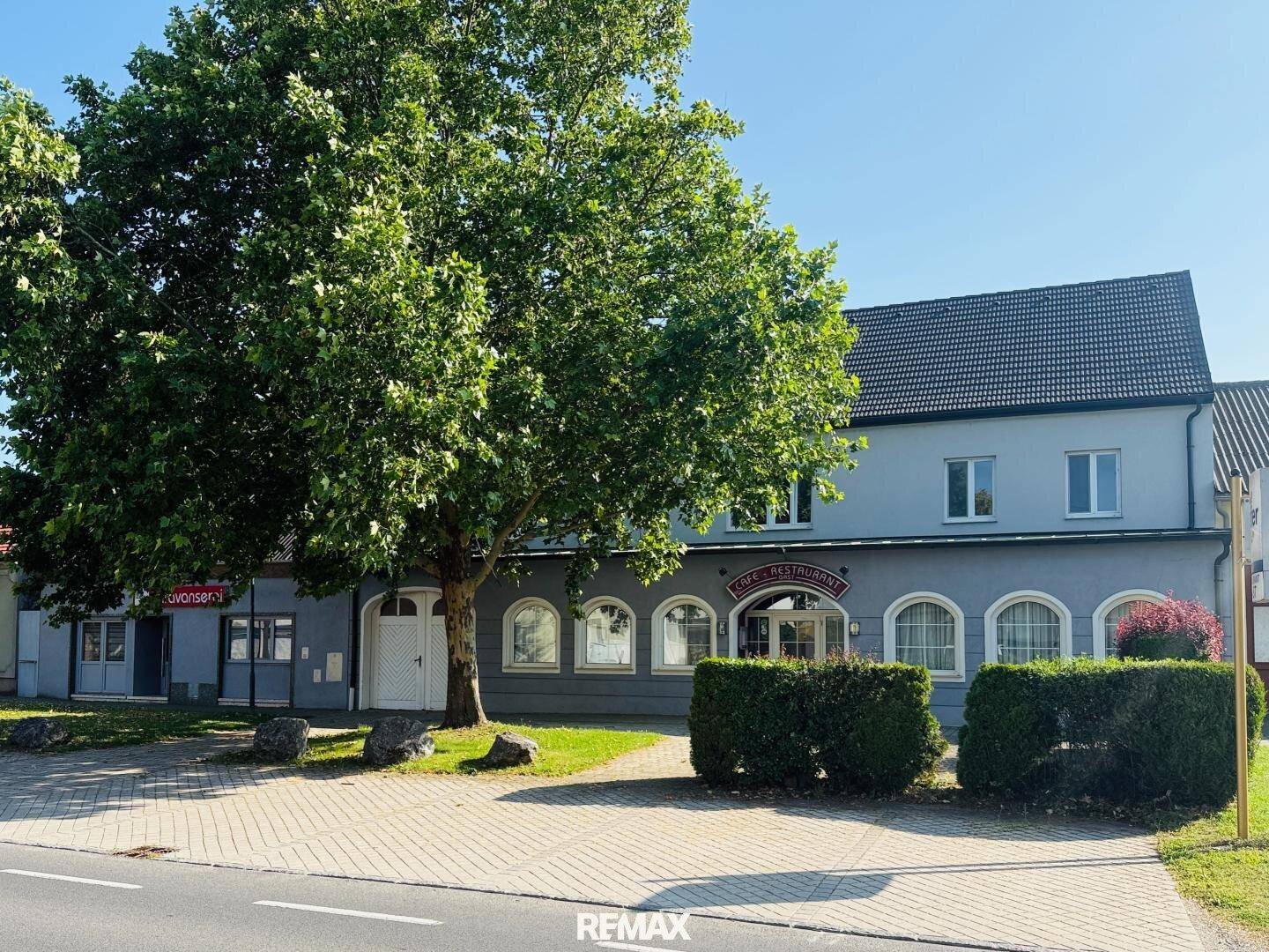 Straßenansicht eines Gebäudes mit "Cafe Restaurant"-Schild, großem Baum und gepflegter Hecke.