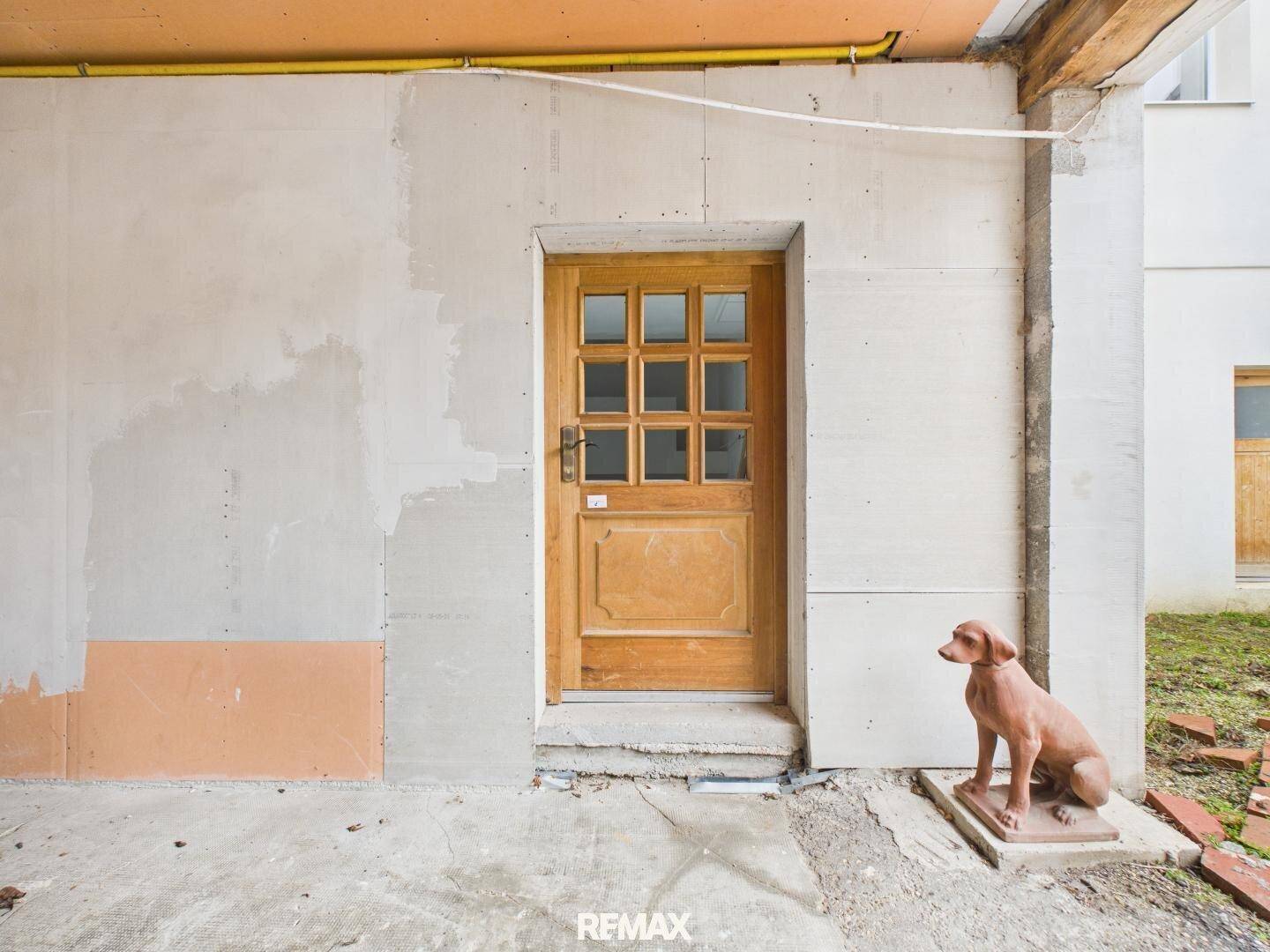 Unfertiger Eingangsbereich mit Holztür und Rohbauwänden, ergänzt durch eine dekorative Hundestatue.