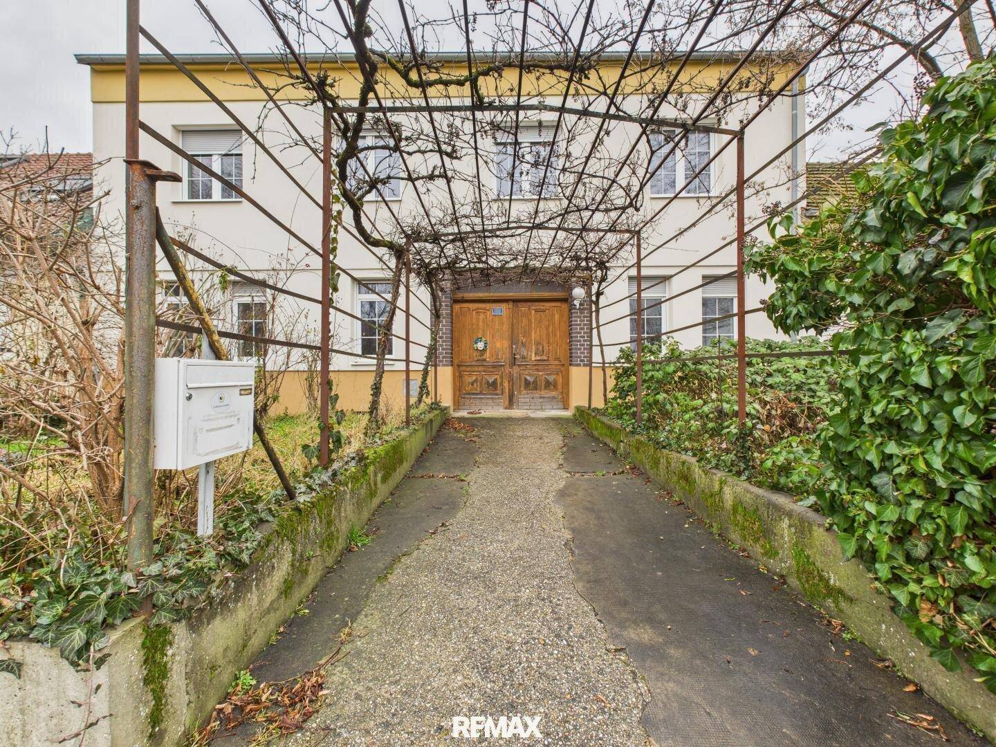 Eingangsbereich eines Hauses mit bewachsenem Vordach und traditionellen Holztüren, umgeben von Vegetation.