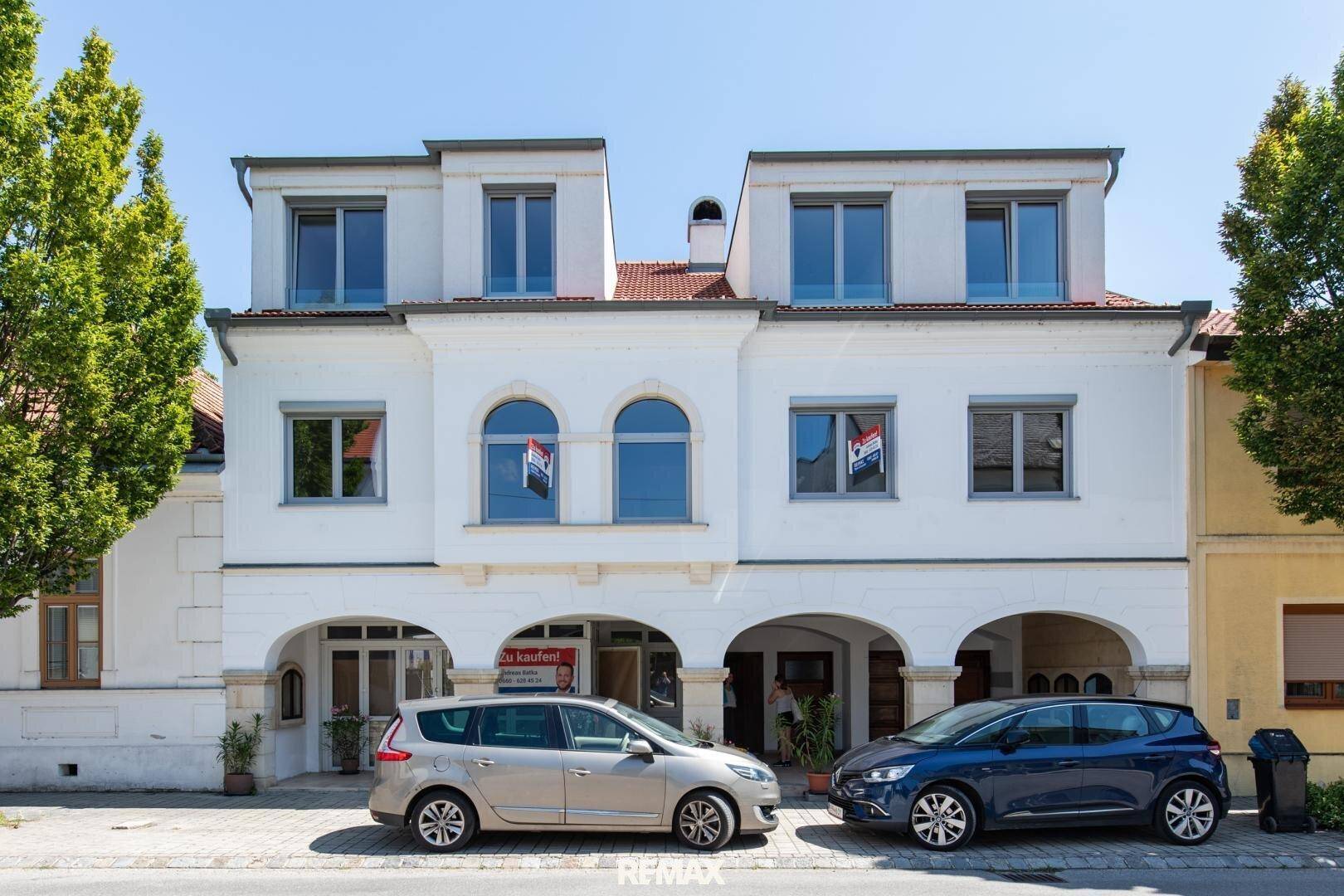 Traditionelles Gebäude mit weißer Fassade, Mansarddach und Arkaden im Erdgeschoss.