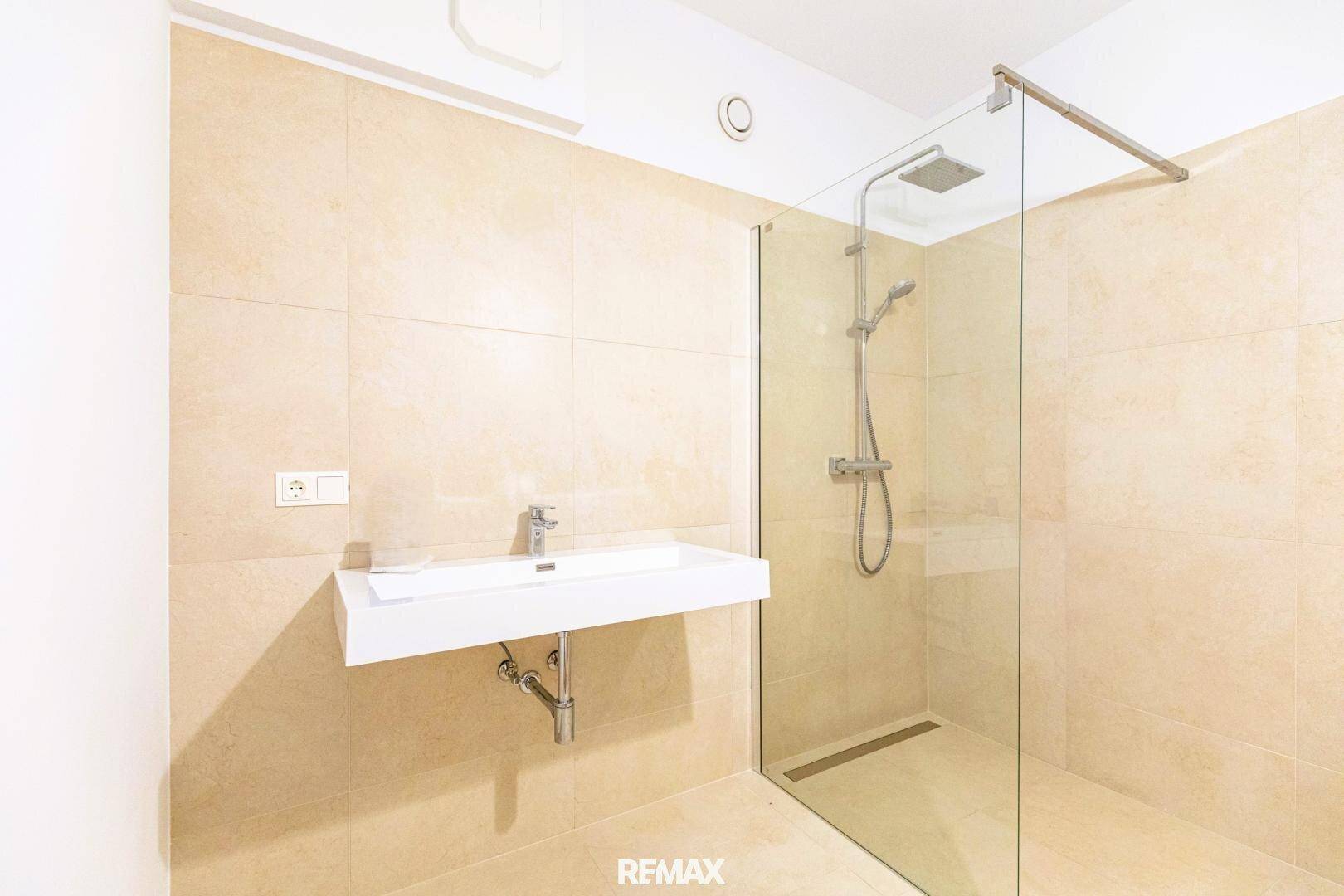 Modernes Badezimmer mit beigefarbenen Fliesen, wandhängendem Waschbecken und begehbarer Dusche.