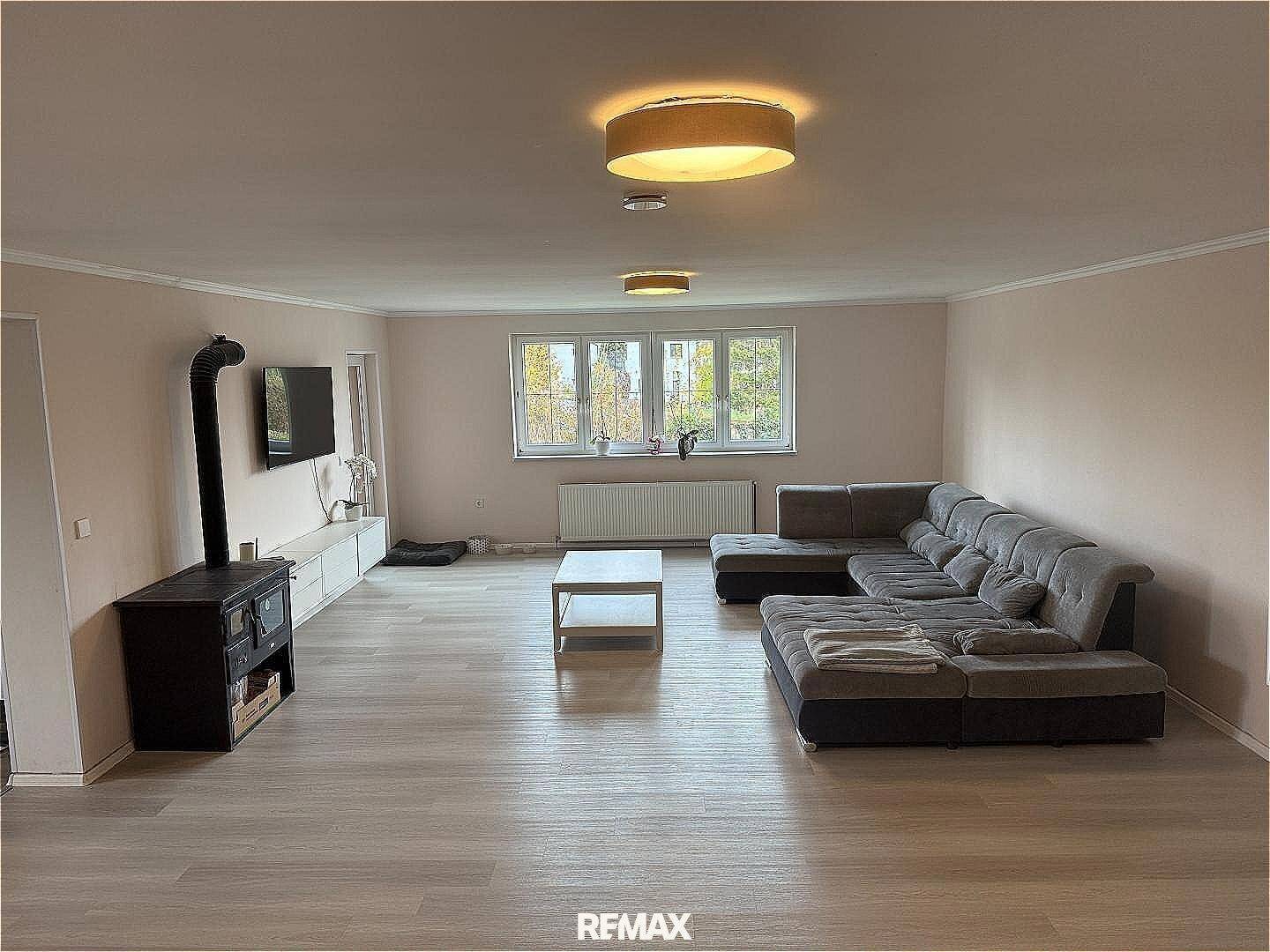 Helles Wohnzimmer mit großem Ecksofa, Kaminofen und Blick ins Grüne durch die Fenster.