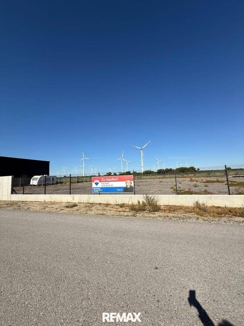 Grundstück mit Zaun, einem Gebäude und Windkraftanlagen in der Ferne unter strahlend blauem Himmel.