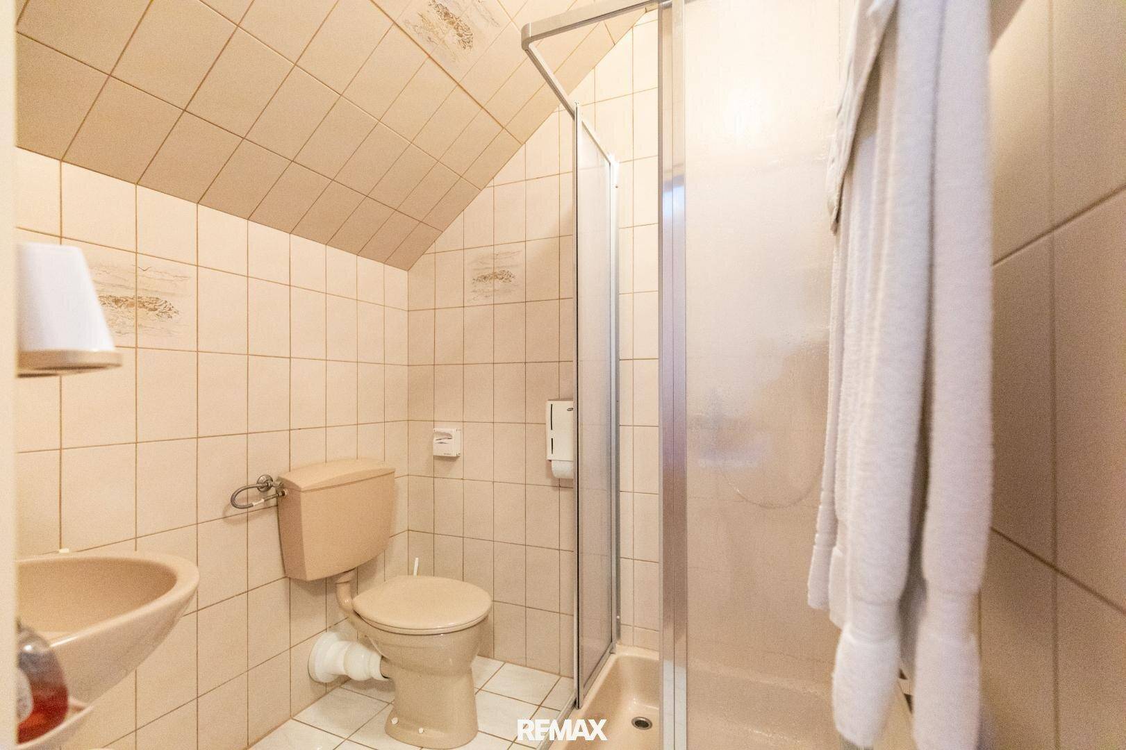 Kleines Badezimmer mit Dachschräge, Duschkabine, WC und Waschbecken, komplett gefliest.