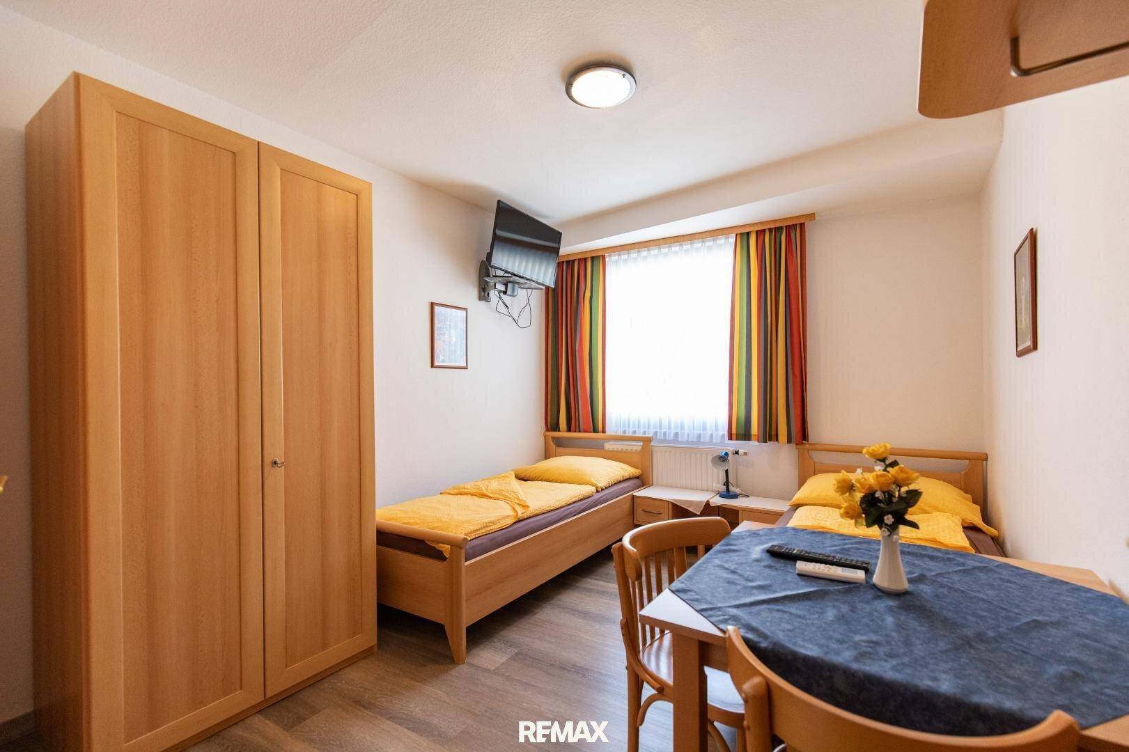 Gemütliches Schlafzimmer mit zwei Einzelbetten, Holzschrank, Tisch und einem Fenster mit Vorhängen.