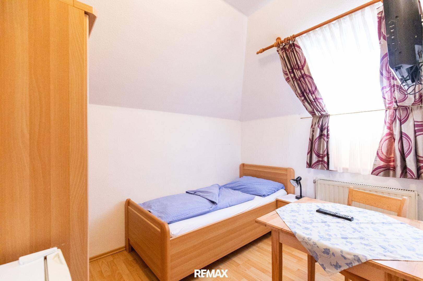 Schlafzimmer mit Einzelbett, Holzboden, Tisch und einem Fenster mit gemusterten Vorhängen.