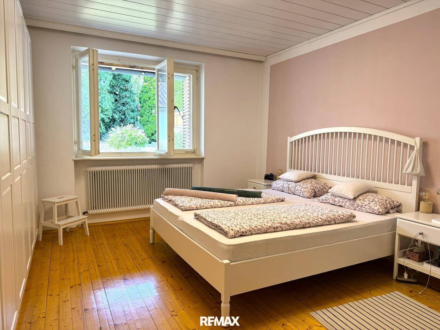 Helles Schlafzimmer mit Holzboden, weißem Doppelbett und Blick ins Grüne durch das Fenster.