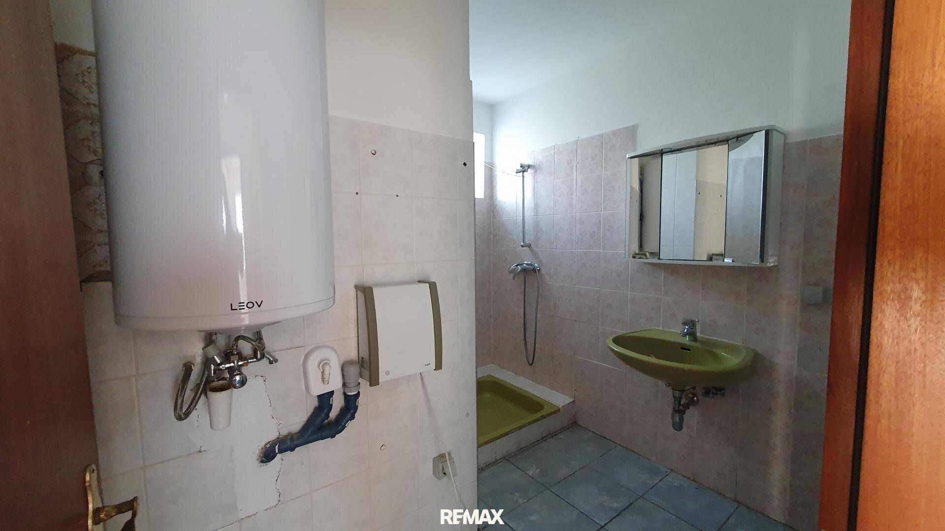 Badezimmer in Originalzustand mit älteren Fliesen, Boiler, Waschbecken und einfacher Dusche.