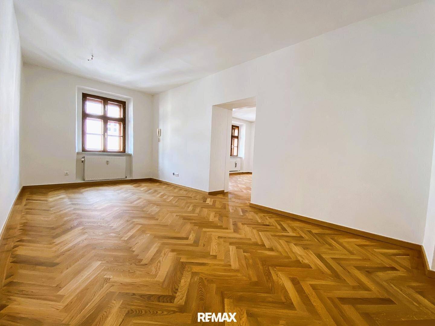 Großer, heller Wohnbereich mit elegantem Fischgrätparkett und Blick in einen angrenzenden Raum.