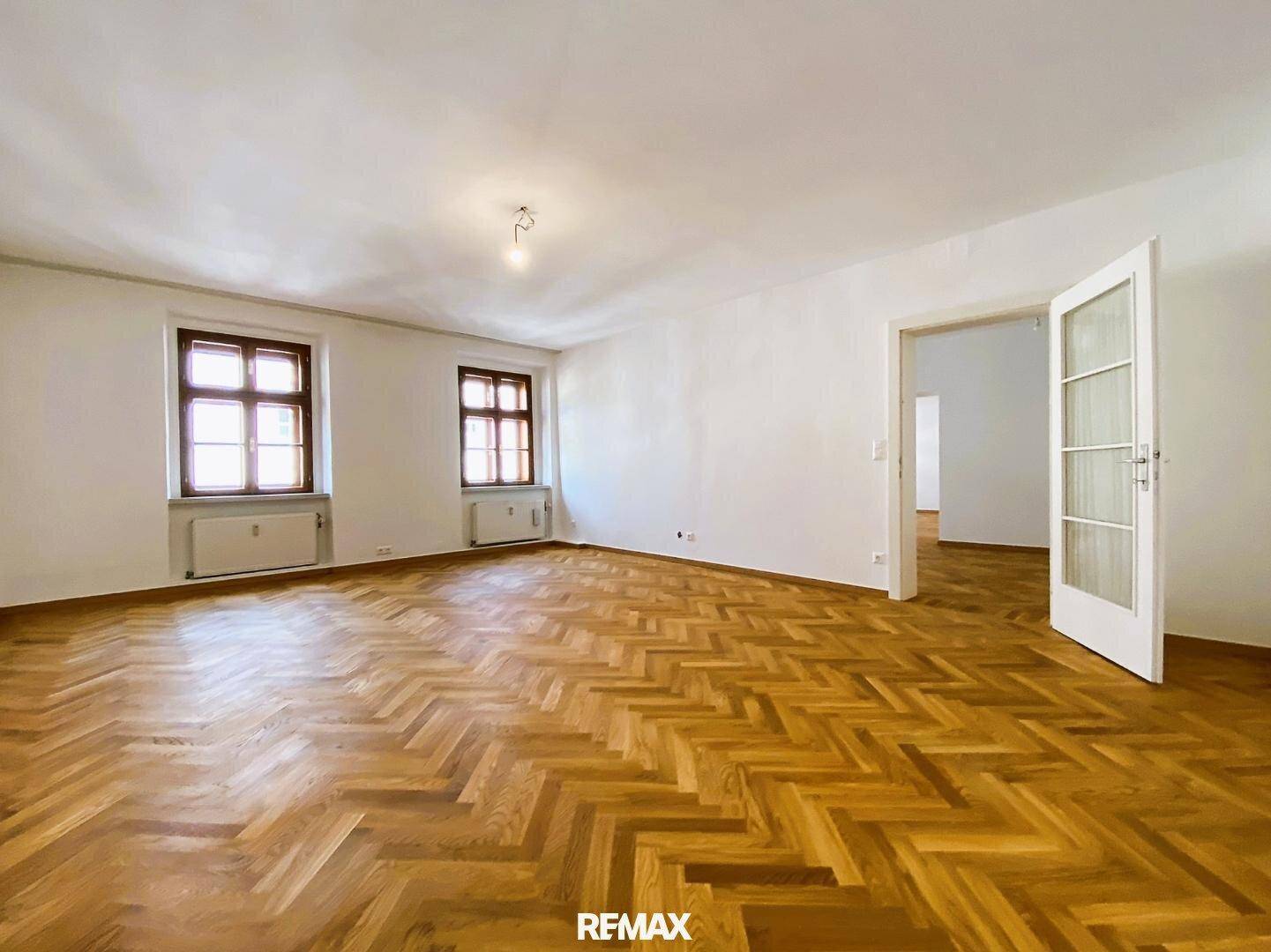 Heller Wohnraum mit klassischem Fischgrätparkett und einer Tür, die zu einem weiteren Zimmer führt.