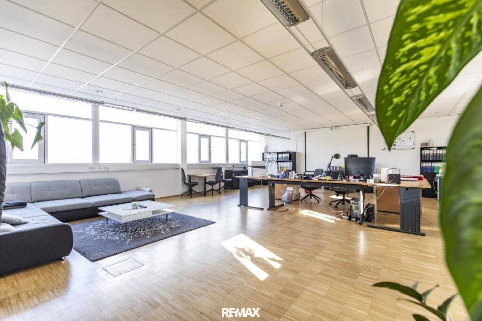 Weitläufiges Großraumbüro mit vielen Fenstern, Schreibtischen und einem gemütlichen Sofa.