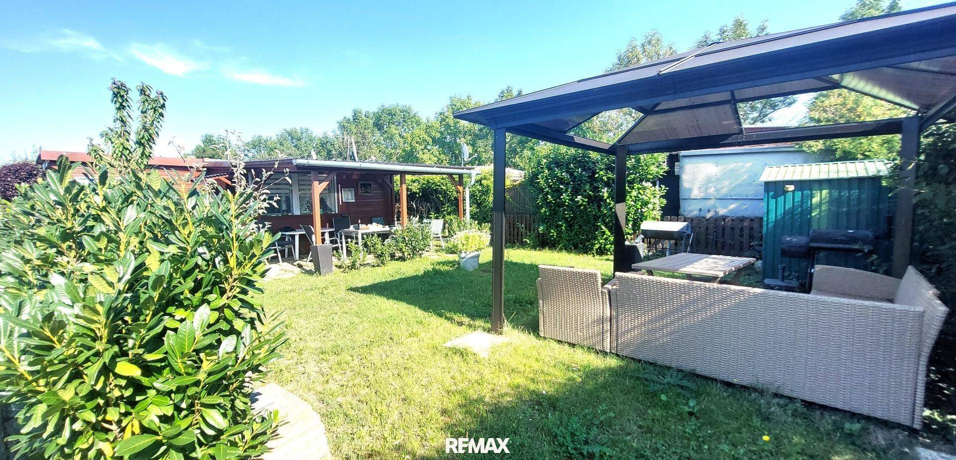Sonniger Gartenbereich mit Rasen, einer gemütlichen überdachten Sitzecke und einem Geräteschuppen.