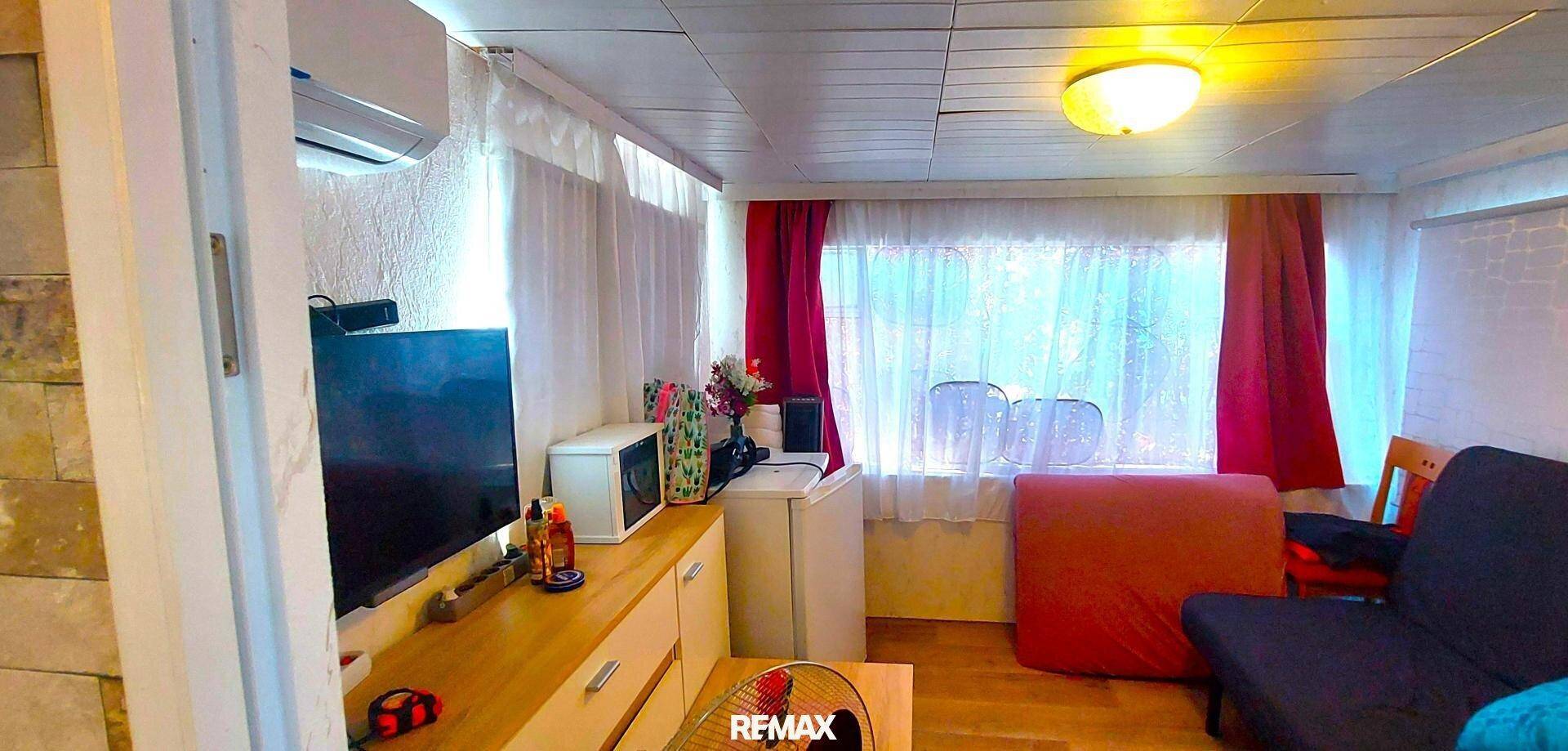 Gemütlicher Wohnbereich mit Fernseher, Sofa und Klimaanlage, beleuchtet durch ein Fenster mit roten Vorhängen.