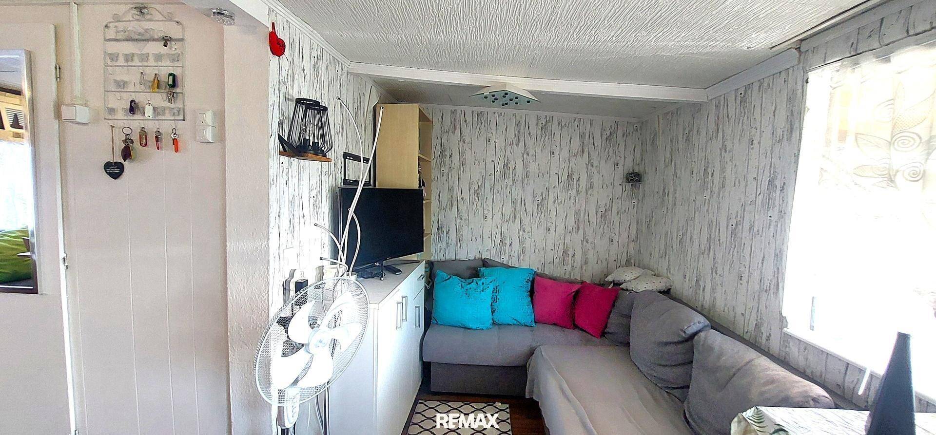 Wohnraum mit Ecksofa, Fernseher und Ventilator, die Wände sind mit heller Holzoptik-Tapete verkleidet.