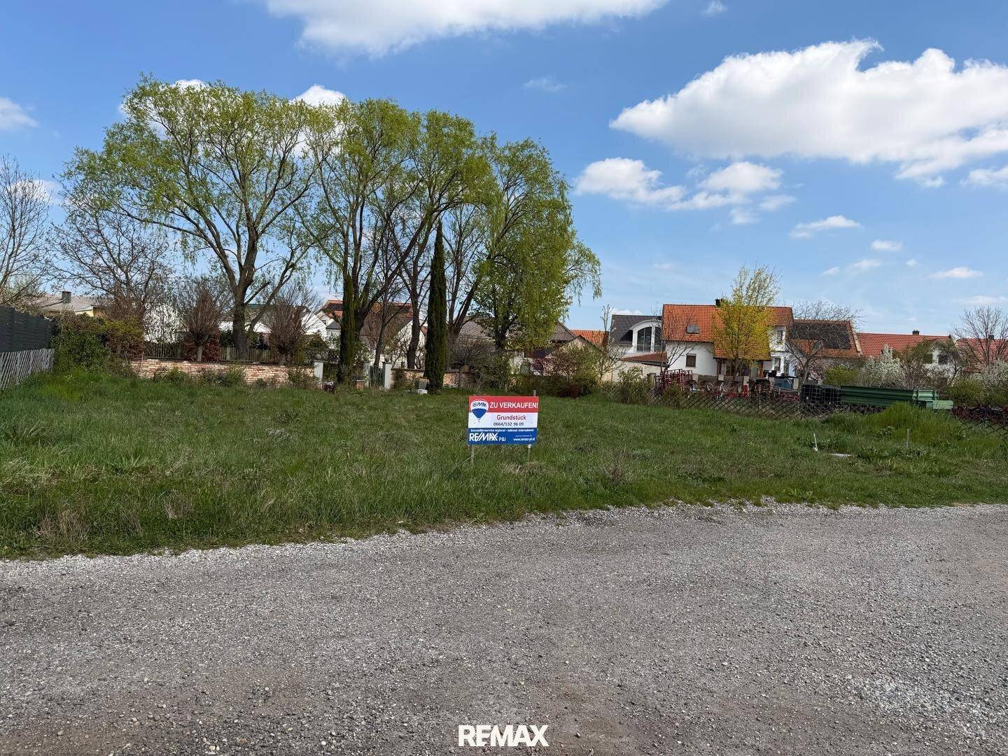 Freies Baugrundstück mit einem "Zu verkaufen"-Schild und Bäumen im Hintergrund unter blauem Himmel.