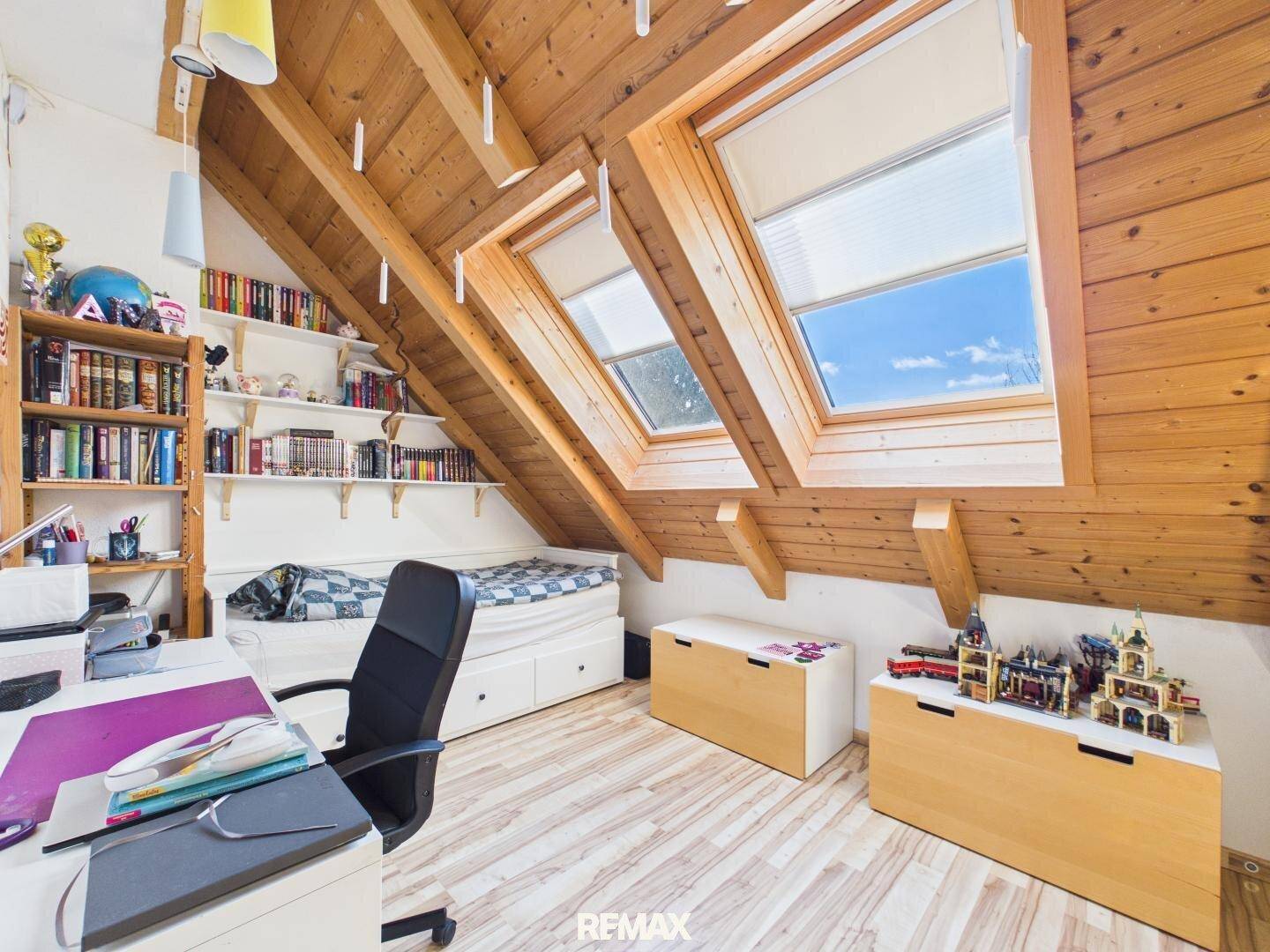 Dachgeschosszimmer mit Holzbalken, Bücherregalen, Bett und zwei großen Dachfenstern.