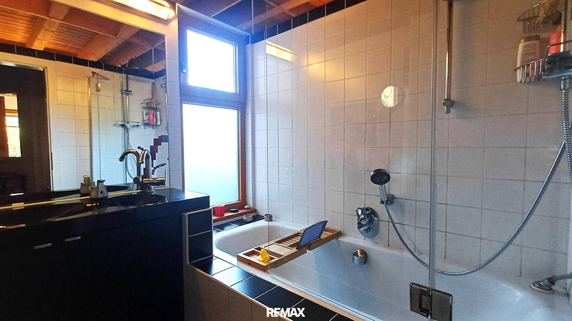 Badezimmer mit Badewanne, Duschbereich, Fenster und dunklen Fliesenakzenten.