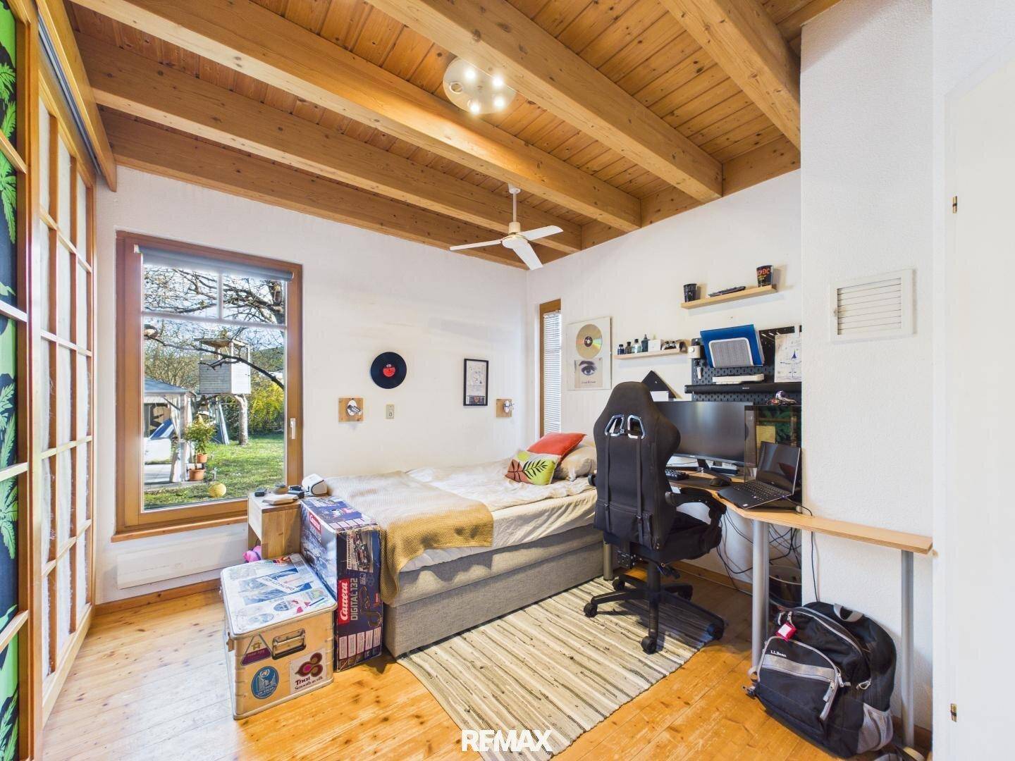 Schlafzimmer mit Holzbalkendecke, großem Fenster zum Garten und integriertem Arbeitsbereich.
