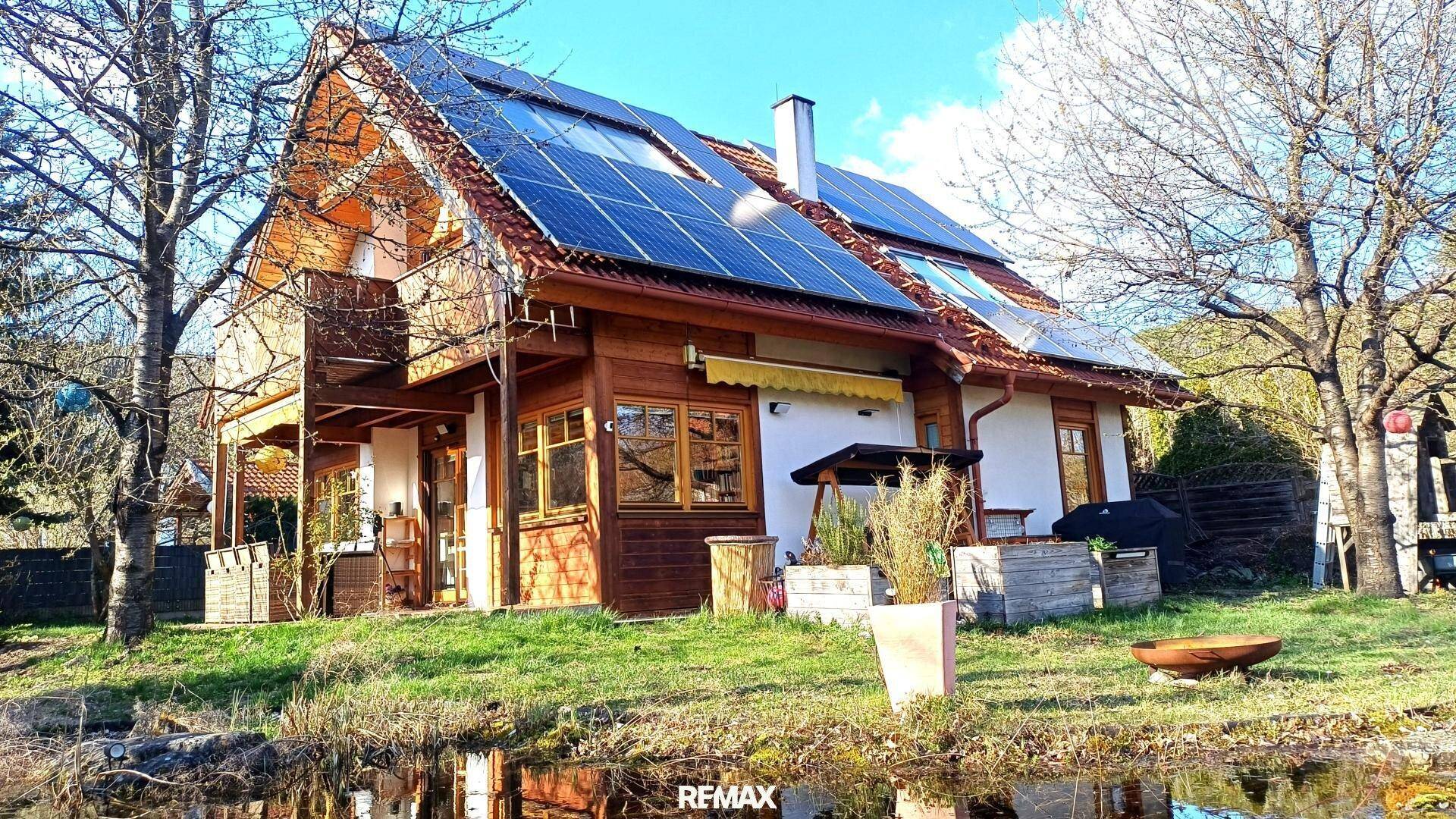 Holzhaus mit Solaranlage auf dem Dach, umgeben von Bäumen und einem spiegelnden Teich.