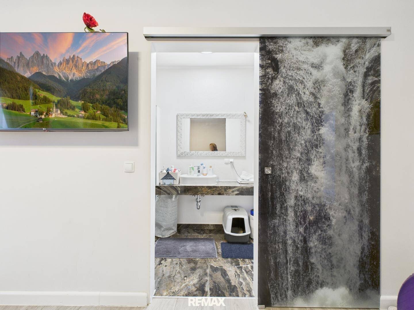 Kleiner Toilettenbereich mit Waschbecken, hinter einer Schiebetür mit Wasserfallmotiv.