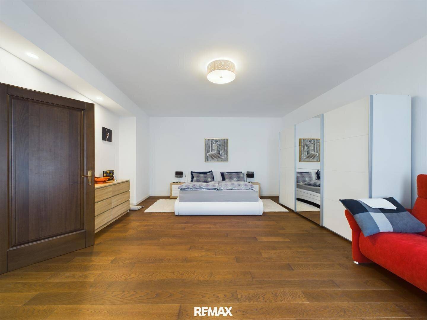 Helles Schlafzimmer mit Holzboden, großem Bett, weißen Schränken und einem roten Sofa.