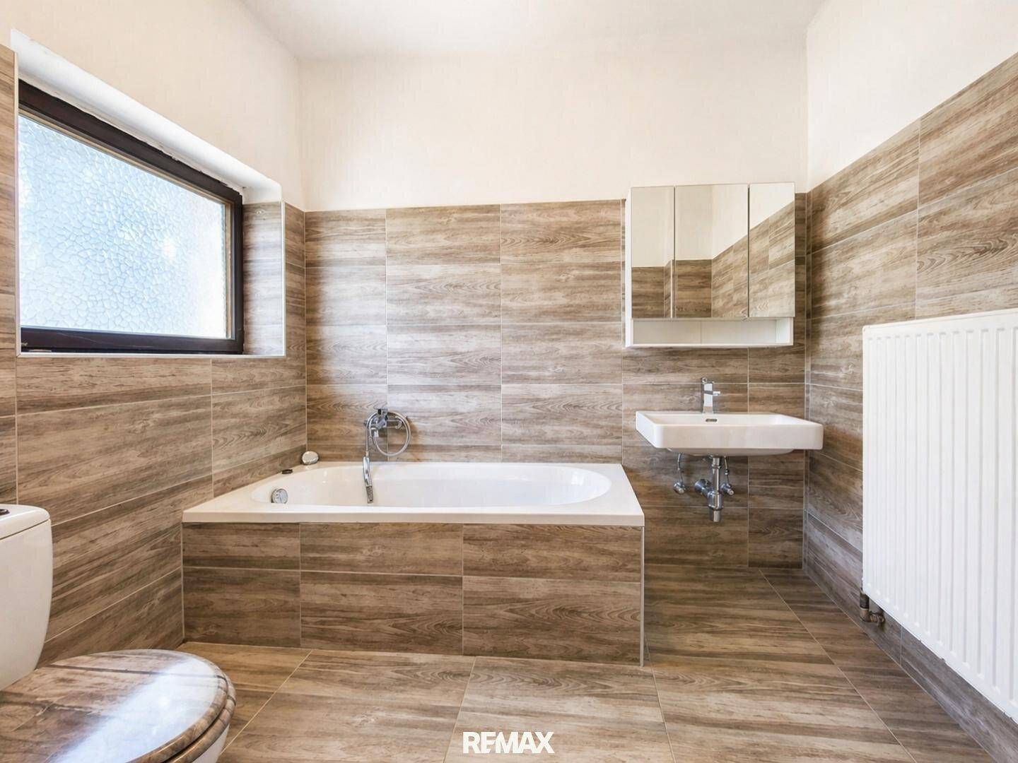 Modernes Badezimmer mit Badewanne, Waschbecken, Spiegelschrank und holzähnlichen Fliesen.