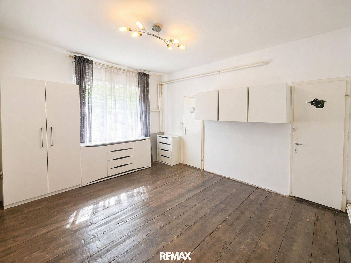 Schlafzimmer mit Holzdielenboden, weißem Schrank und großem Fenster.
