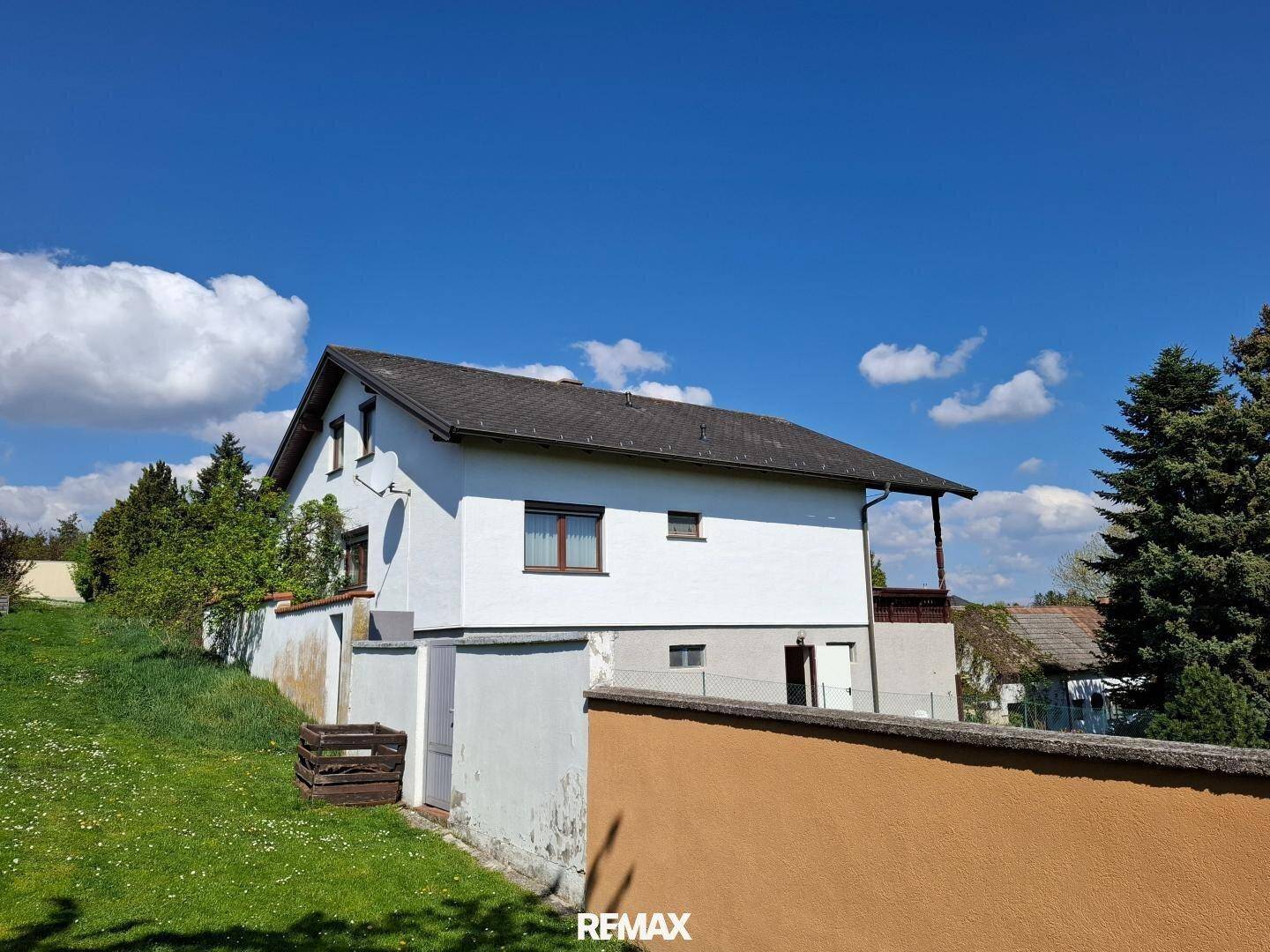 Weißes Einfamilienhaus mit dunklem Dach und umzäuntem Grundstück unter blauem Himmel.