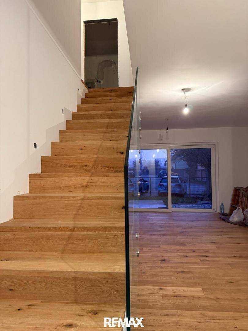 Moderne Holztreppe mit Glasgeländer, die zu einem hellen Wohnbereich mit großen Fenstern führt.