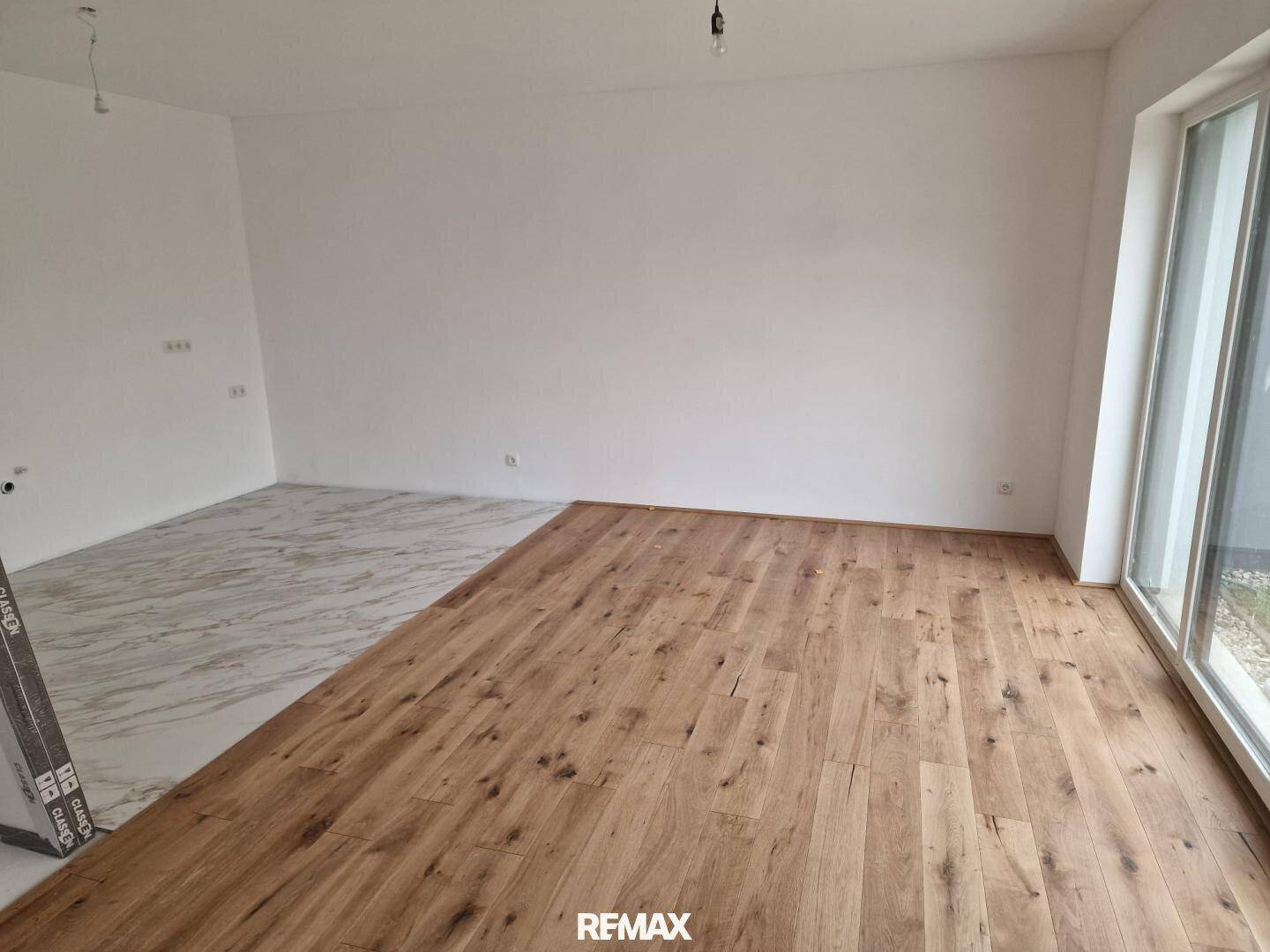 Geräumiger Raum mit teilweise verlegtem Holz- und Fliesenboden, großen Fenstern und viel Tageslicht.