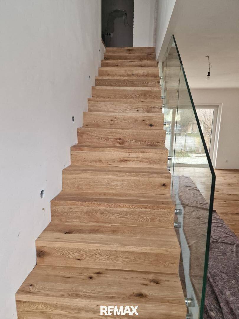 Moderne Holztreppe mit Glasgeländer in einem hellen Innenraum, der noch im Bau ist.