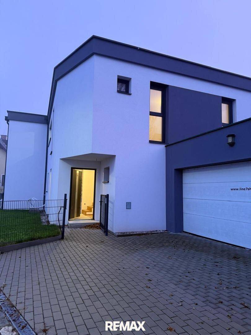 Modernes Haus bei Dämmerung mit beleuchteten Innenräumen und integrierter Garage.