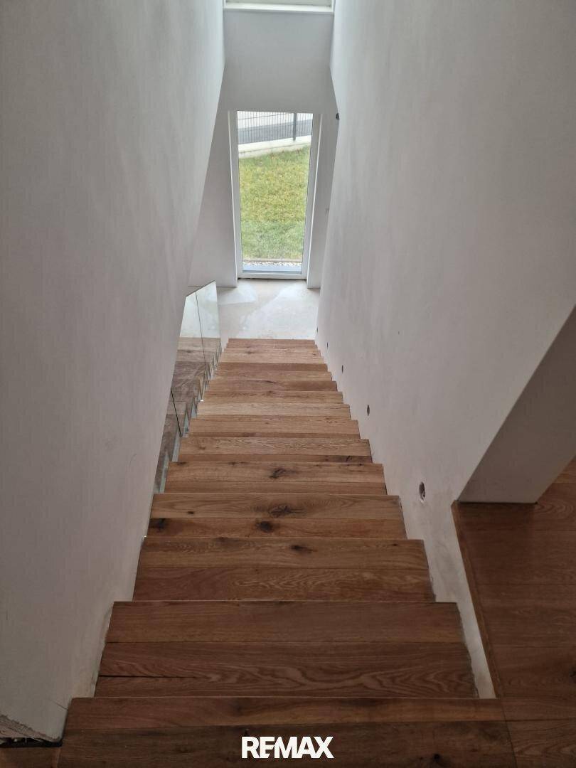 Moderne Holztreppe mit Glasgeländer und Blick auf den Außenbereich durch die Tür am unteren Ende.
