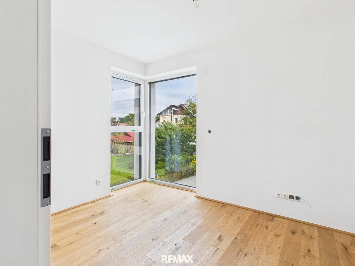 Helles Zimmer mit Holzboden und Eckfenstern, die einen Blick ins Grüne bieten.
