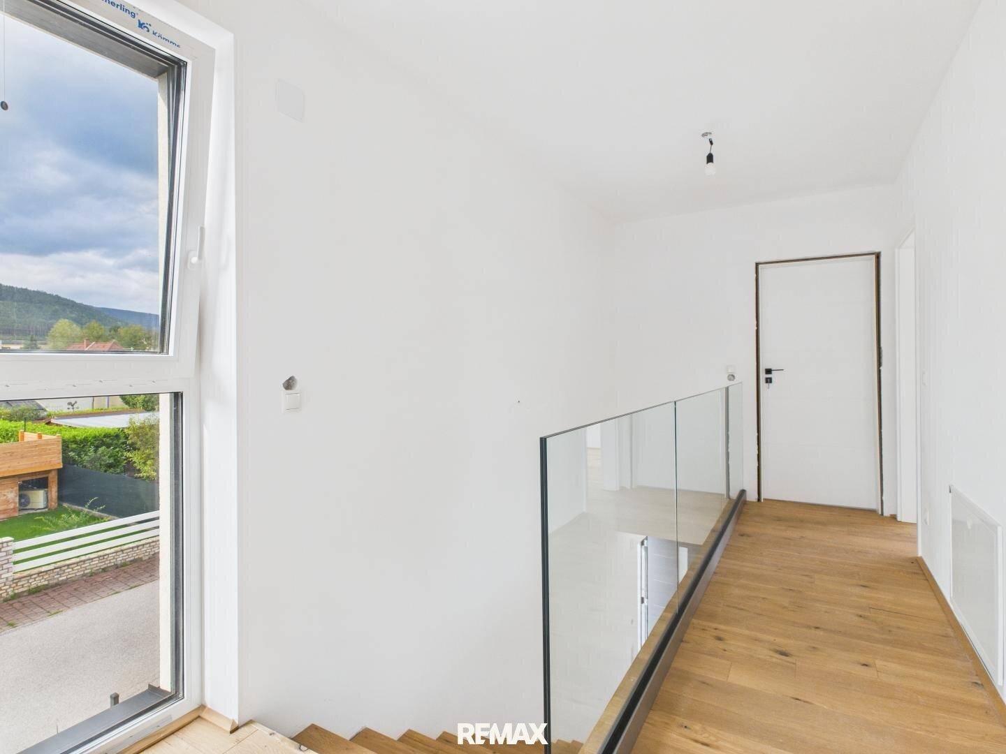 Heller Flur im Obergeschoss mit Holzfußboden, Glasgeländer und großem Fenster.