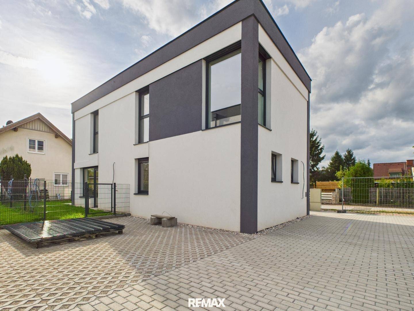 Moderne Architektur des Neubaus mit weißer und grauer Fassade und gepflastertem Vorplatz.